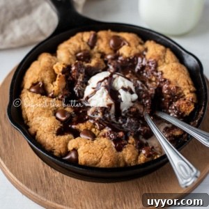 Warm chocolate chip skillet cookie with scoop of vanilla ice cream and two spoons resting in skillet on wood trivet.
