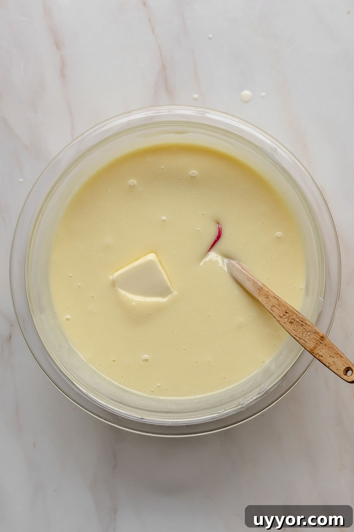 A pat of butter in the white chocolate ganache with a spatula resting in the bowl.