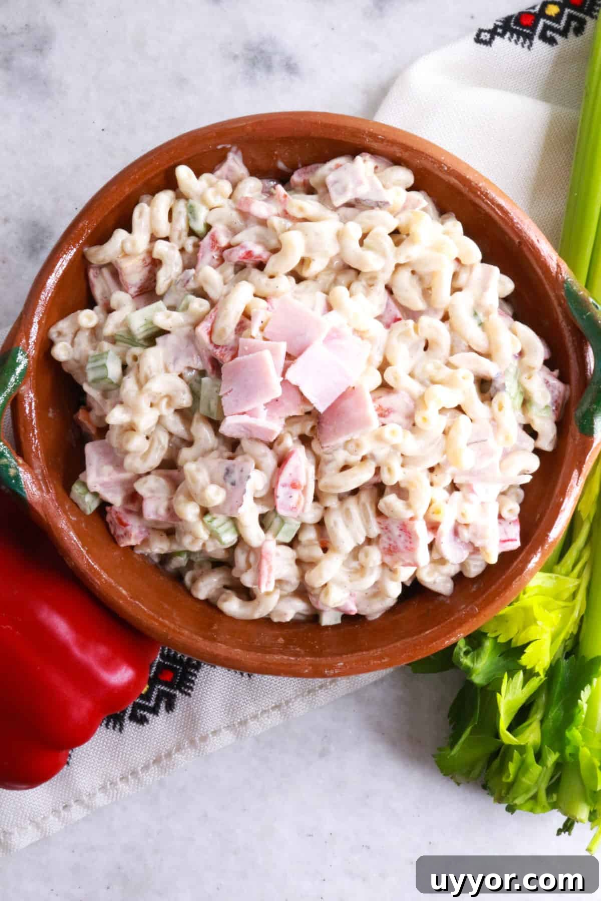 Ensalada de Coditos in a brown bowl with a red bell pepper and stalk of celery beside it.