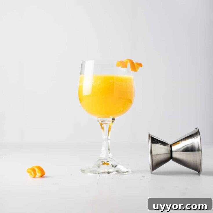 A pineapple old fashioned cocktail with an orange peel garnish next to a toppled jigger and extra orange garnish