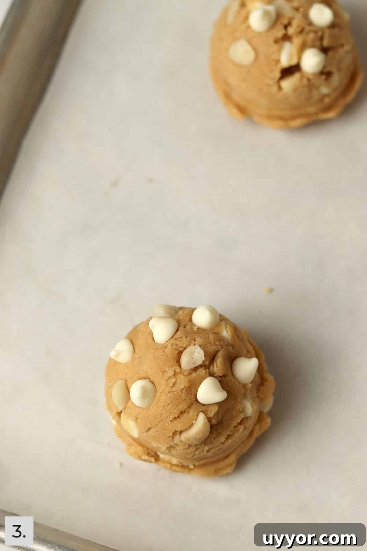 Two white chocolate macadamia nut cookie dough balls on a baking sheet.