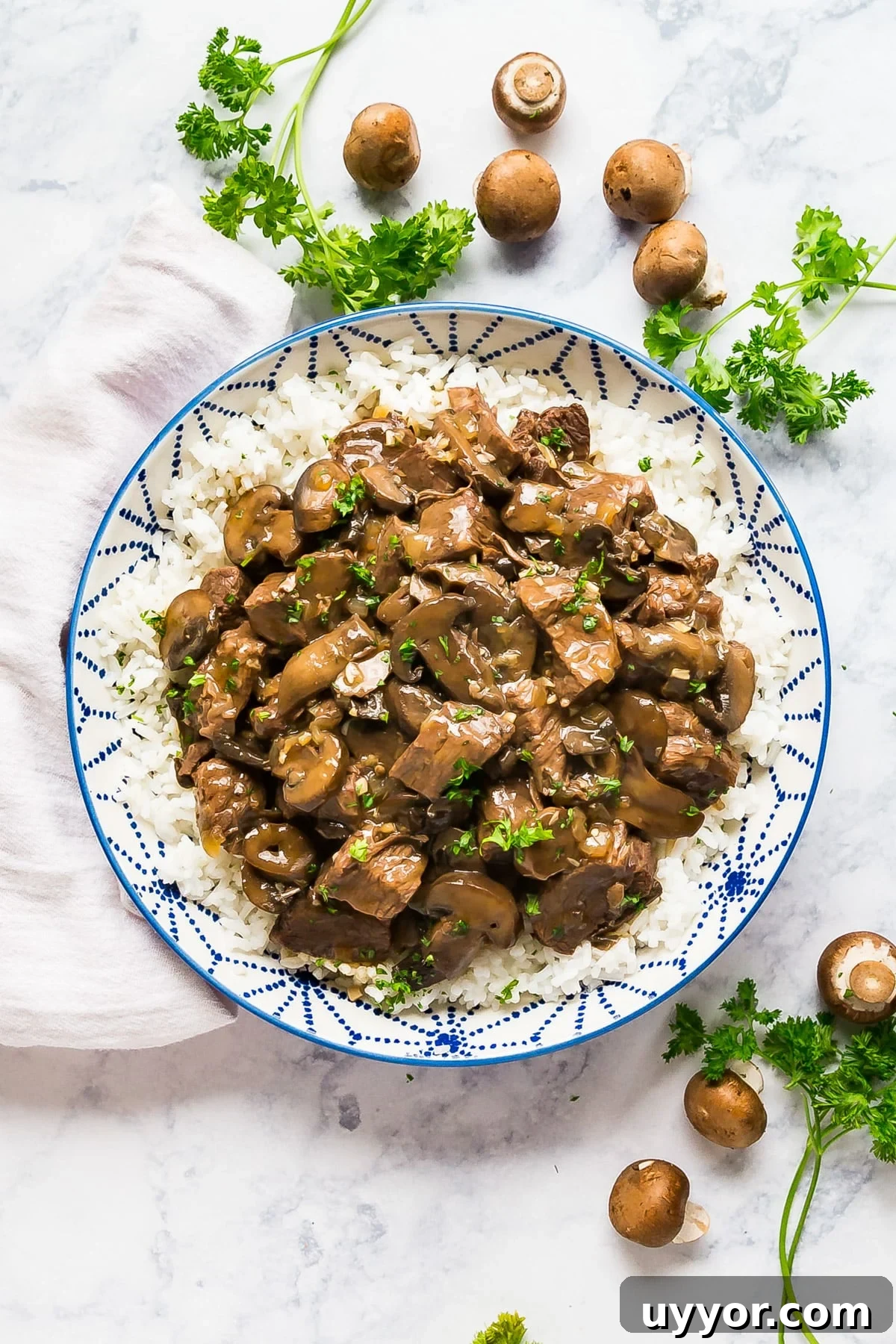 Instant Pot Beef Tips with Rich Mushroom Gravy 2 An overhead image of a blue and white bowl of beef tips over rice with parsley and mushrooms around it.