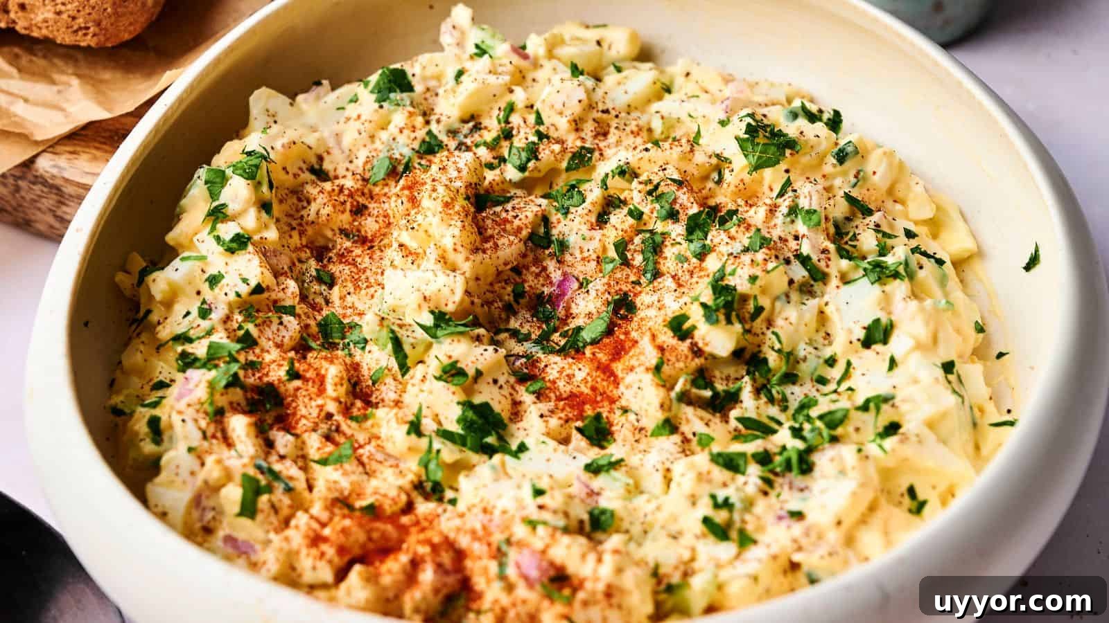 A bowl of creamy egg salad topped with chopped parsley and paprika, set on a light-colored surface.