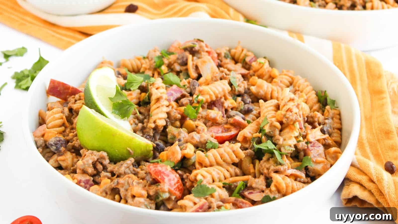 A bowl of fusilli pasta mixed with ground beef, black beans, chopped tomatoes, and herbs, garnished with lime wedges.