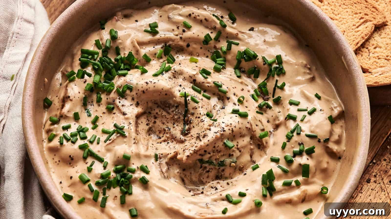 Bowl of creamy dip topped with chopped chives and black pepper, next to pieces of toasted bread.