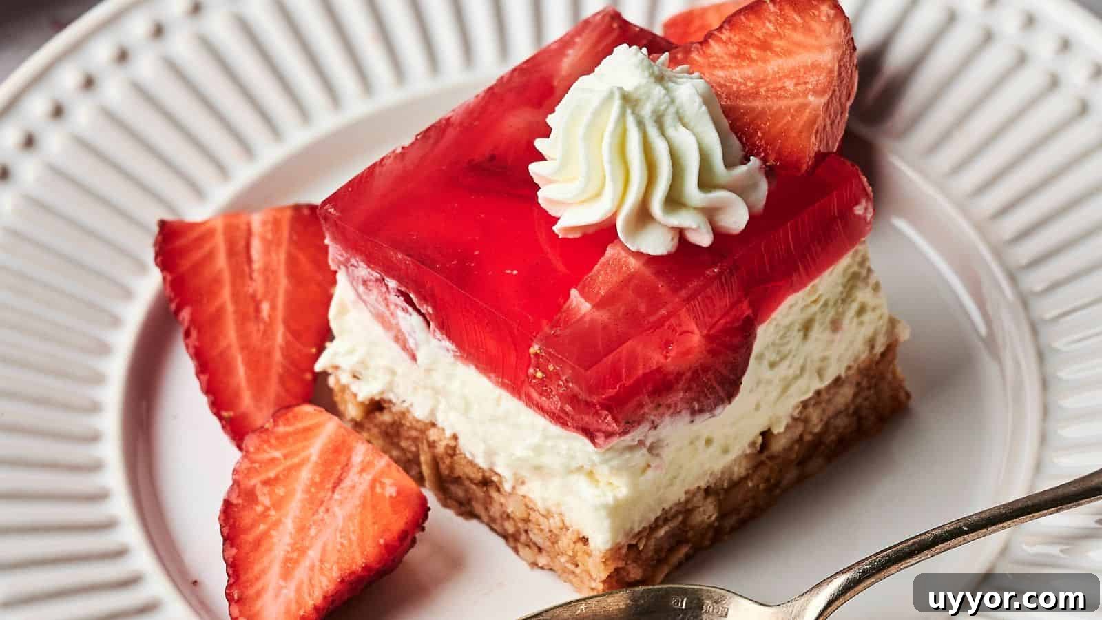 A dessert with a crumbly crust, creamy filling, a layer of red gelatin, topped with a whipped cream dollop and a strawberry slice, on a white plate with a spoon.