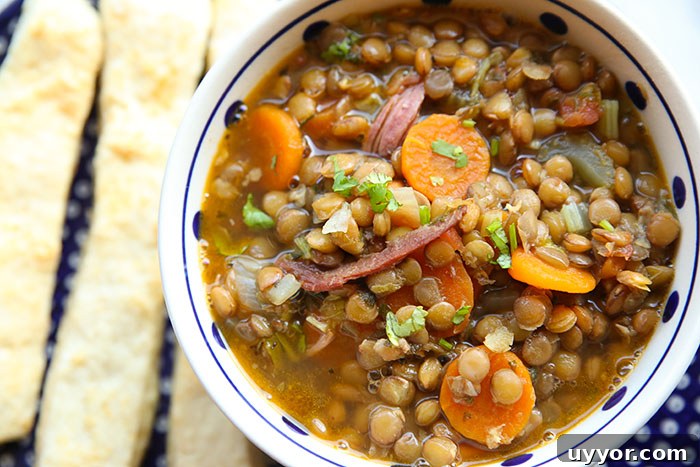 A white and blue polka-dotted bowl filled with ham and lentil soup sitting on a large white polka-dotted blue plate with three bread sticks on the left of the bowl.