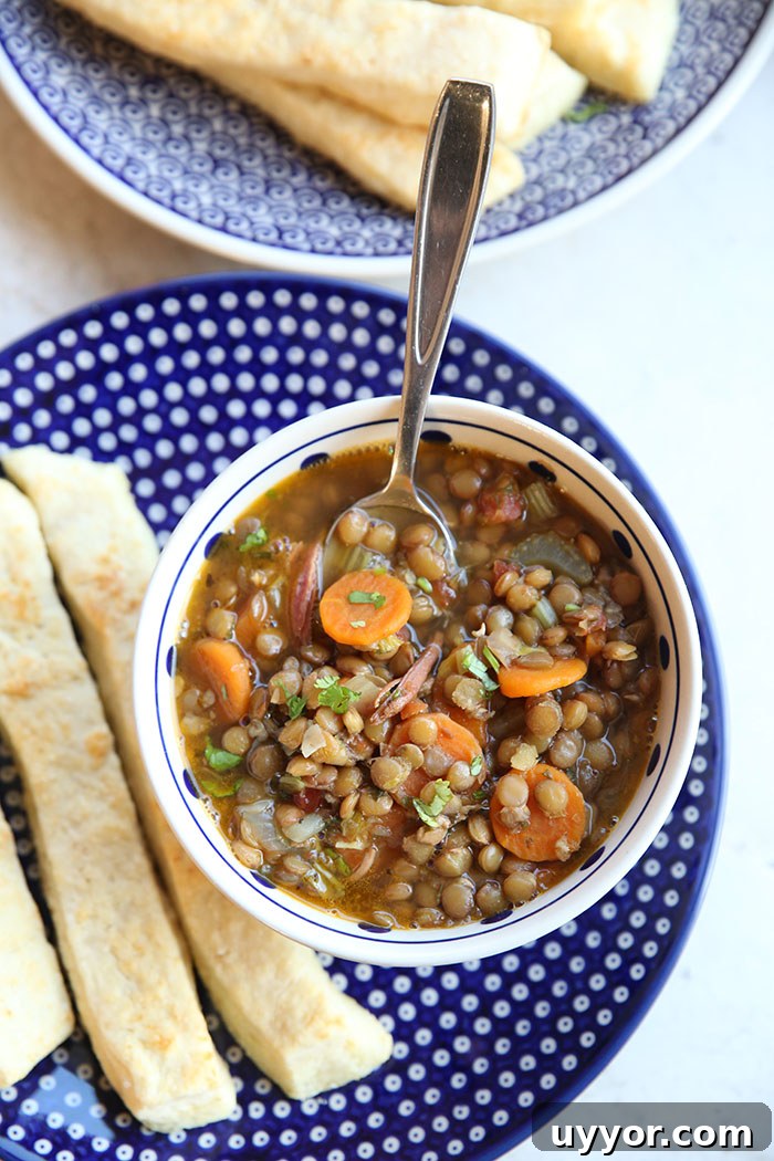 A large dark blue plate with white polka dots with three breadsticks and a blue and white polka-dotted soup bowl of lentil and ham bone soup with a spoon inserted. Next to the plate is a partial picture of a plate of breadsticks.