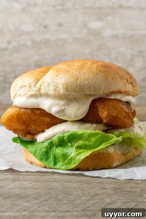Crispy Beer-Battered Cod Sandwich with Tartar Slaw 2 a bun filled with lettuce, white creamy sauce, and golden brown fish filet on parchment paper.