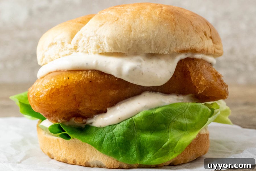 Crispy Beer-Battered Cod Sandwich with Tartar Slaw 10 up close front view of a bun filled with lettuce, white creamy sauce, and golden brown fish filet.