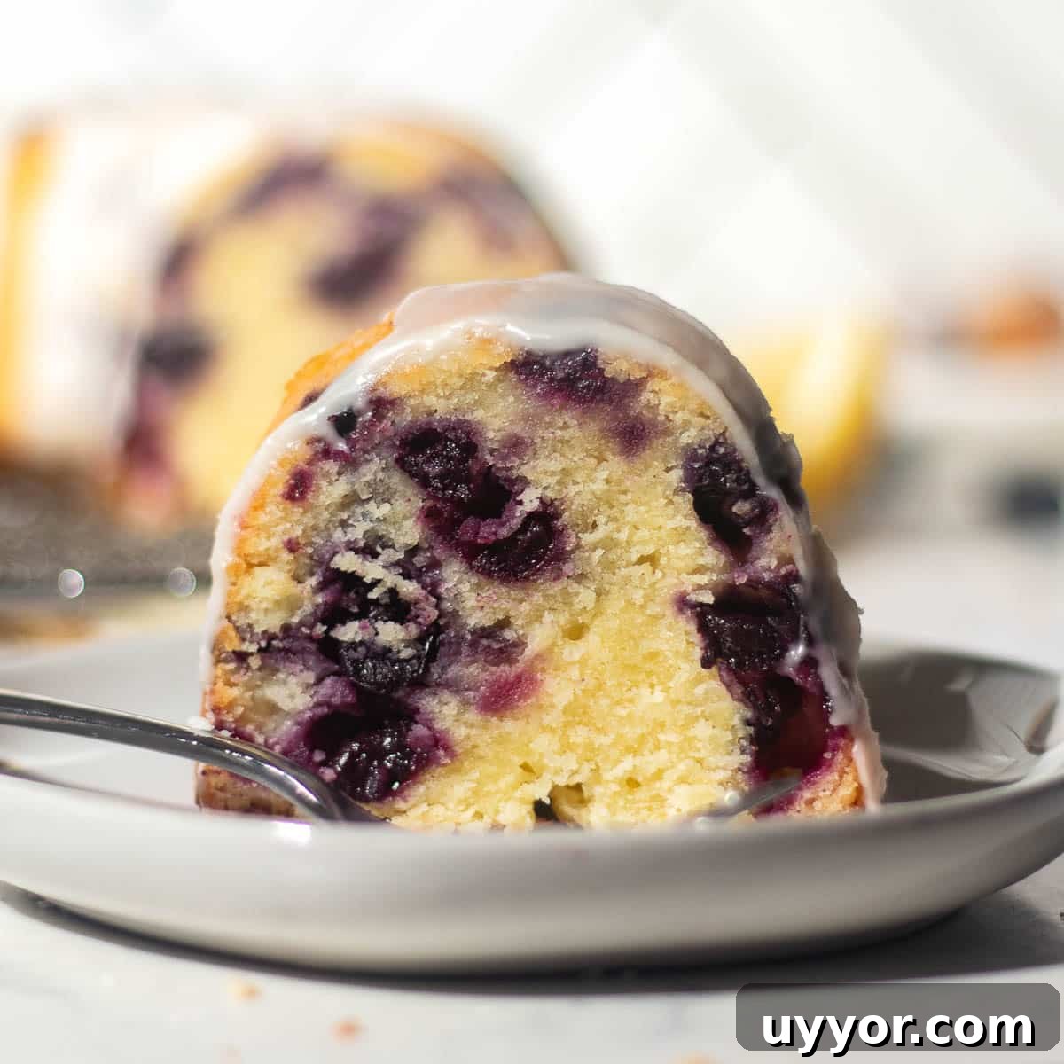 Blueberry Lemon Pound Cake Recipe with Zesty Glaze 3 A slice of blueberry and lemon pound cake on a white plate, with a larger portion of the cake blurred in the background, topped with lemon glaze.