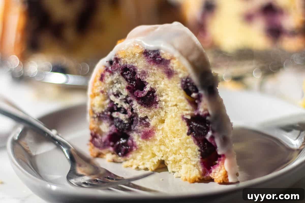 Blueberry Lemon Pound Cake Recipe with Zesty Glaze 6 Close-up of a slice of blueberry and lemon pound cake on a white plate, drizzled with lemon glaze, highlighting the blueberries within the cake.