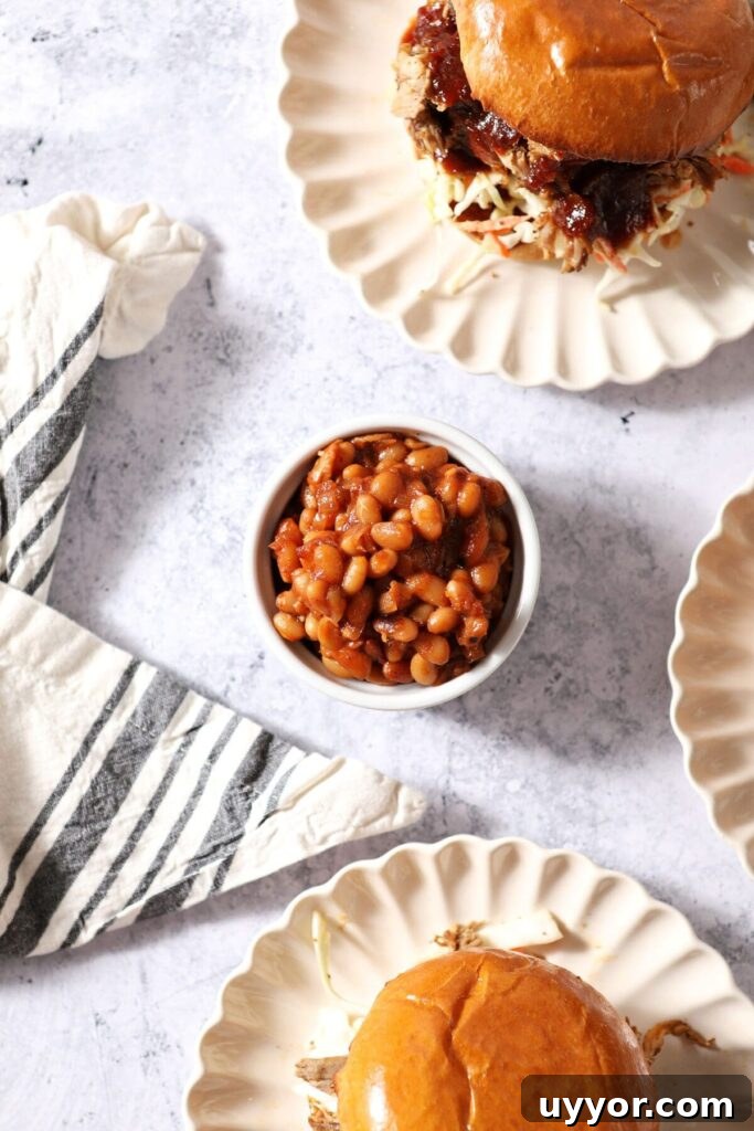 A small ramekin holds a serving of baked beans next to plates of pulled pork sandwiches