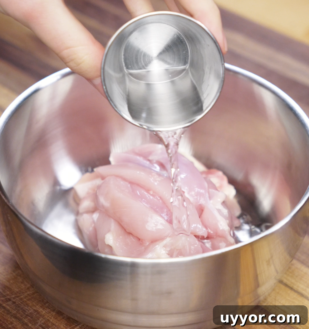 Crispy Pan-Fried Chicken Noodles with Soy-Garlic Sauce 6 A hand pours Shaoxing wine over raw sliced chicken thighs in a metal bowl. This step helps tenderize the meat and infuse it with classic Chinese flavor.