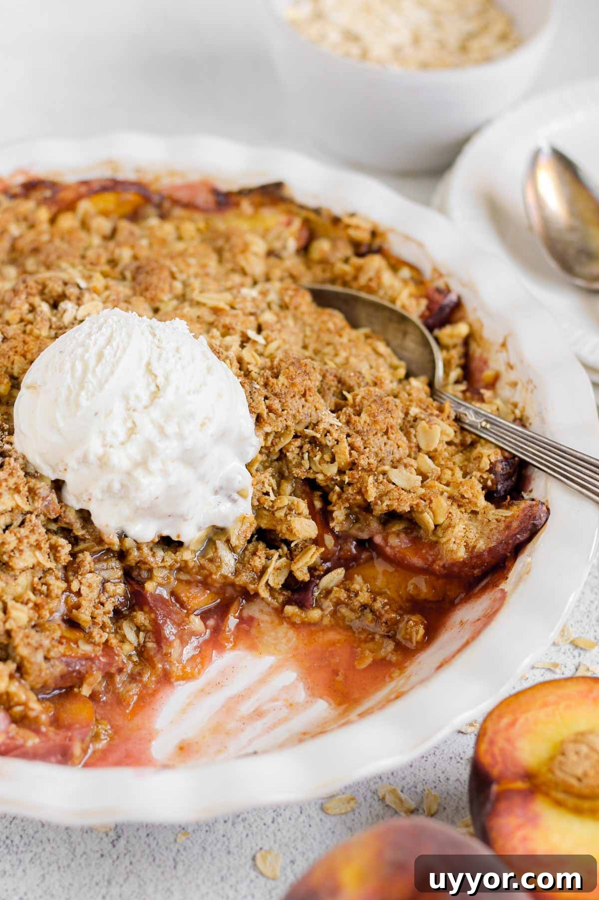 Whole-Grain Peach Crisp with Low Sugar Recipe 11 A baked peach crisp in a white baking dish topped with a scoop of vanilla ice cream. A serving has been scooped out, revealing juicy peaches beneath a golden oat crumble topping. Fresh peach halves and scattered oats sit nearby.