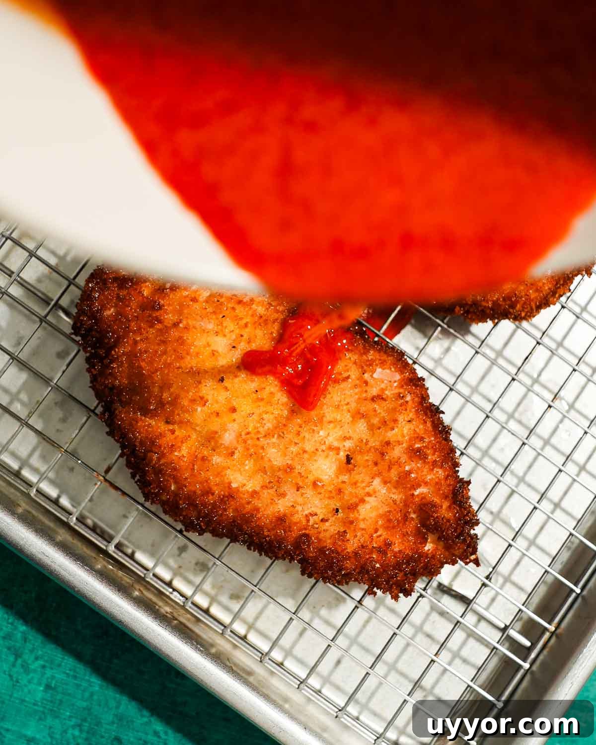 Crispy Panko Buffalo Chicken Cutlets With Tangy Sauce 7 Buffalo sauce being poured onto crispy fried chicken cutlet on rack.