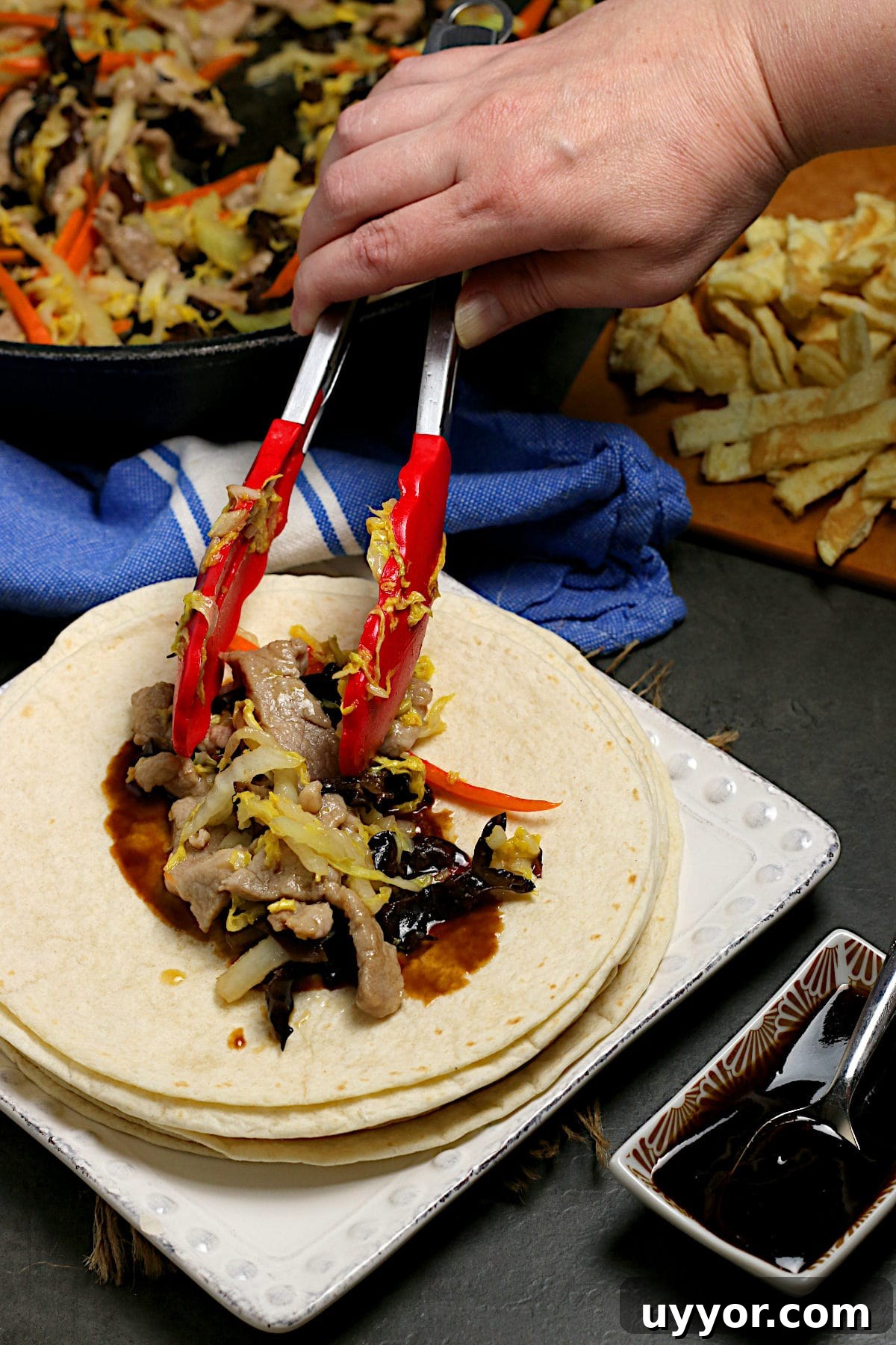 Weeknight Moo Shu Pork Recipe with Quick Hoisin Wraps 4 A hand scooping Easy Weeknight Moo Sho Pork onto a white flour tortilla with tongs.