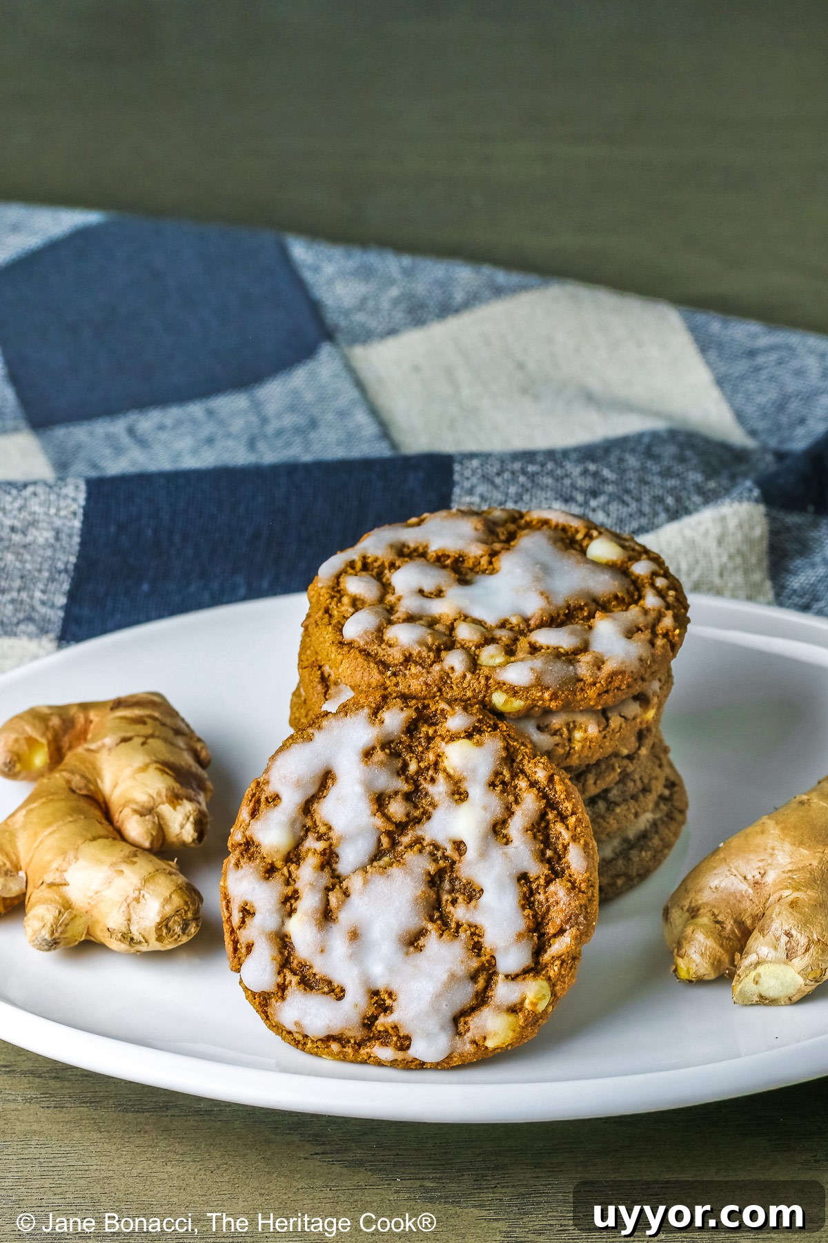 Stack of Iced Ginger White Chocolate Cookies on a white platter with a single iced cookie in front leaning against the stack flanked with two pieces of fresh ginger on either side with a checked cloth behind © 2024 Jane Bonacci, The Heritage Cook. 