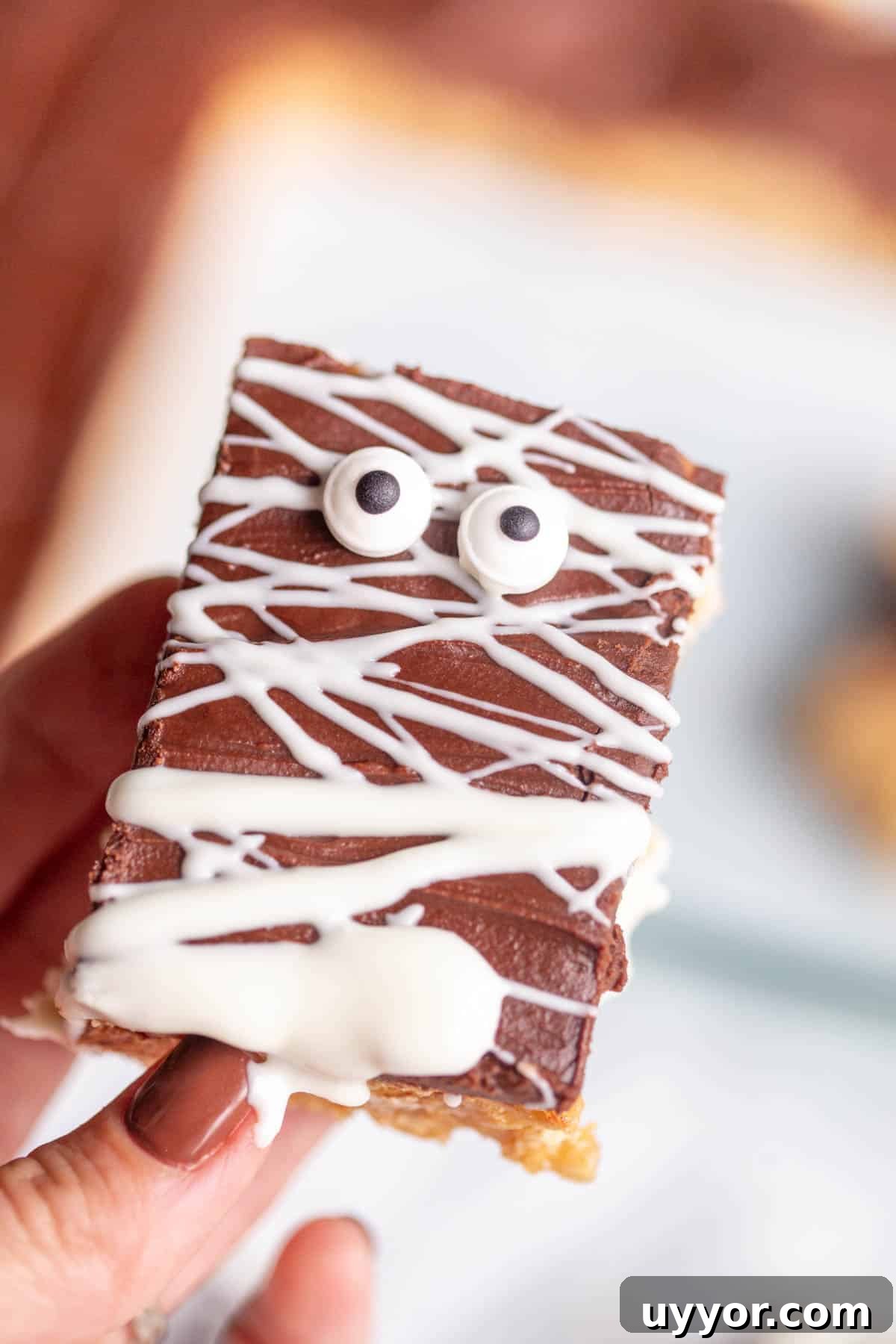 Halloween Scotcheroos Recipe: Spooky No-Bake Peanut Bars 8 A hand with brown finger nails holding a Halloween Scotcharoo drizzled with white chocolate and decorated with edible eyes.