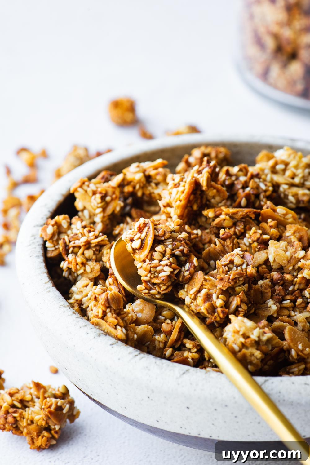 Tahini Sesame Granola with Honey & Orange 3 Close-up of tahini granola with sesame seeds in a ceramic bowl with a jar in the background and a small gold spoon.