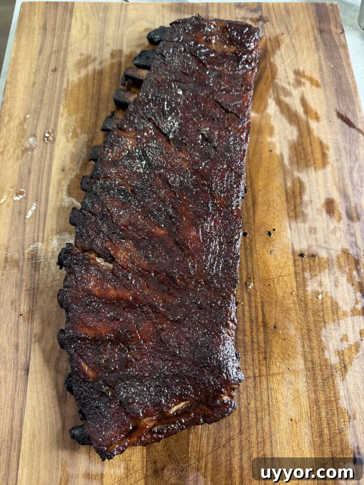 A rack of barbecued ribs with a dark, caramelized glaze rests on a wooden cutting board, showing a smoky, juicy exterior.