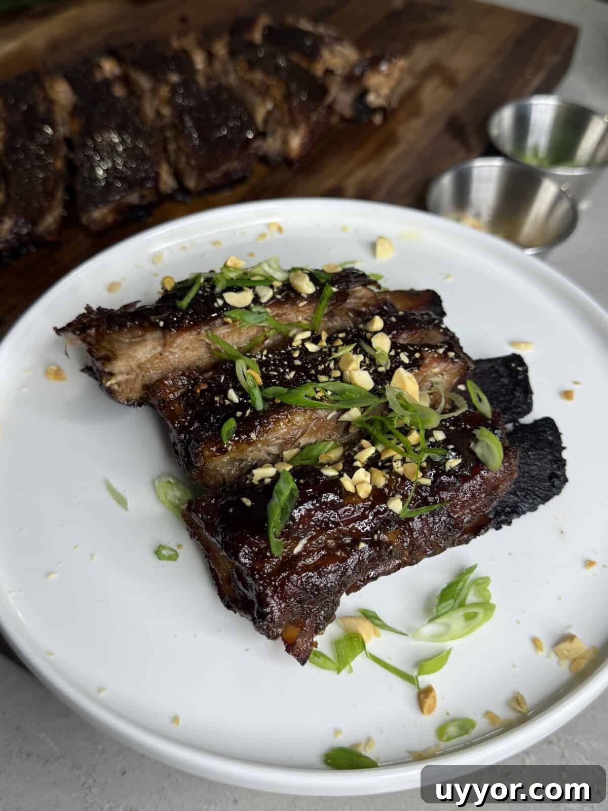 Close-up of glazed pork ribs garnished with chopped green onions and crushed peanuts on a white plate, with more ribs and two metal sauce cups on a wooden board in the background.