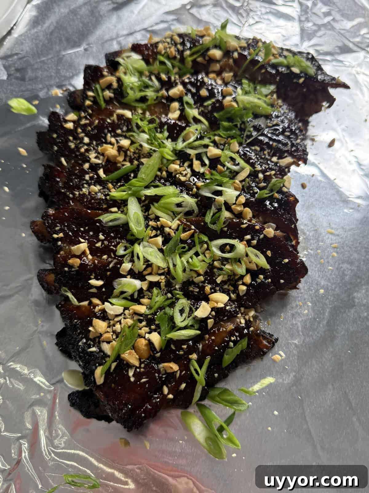 A piece of grilled meat topped with chopped green onions and crushed peanuts, served on a sheet of aluminum foil.