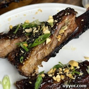 Two glazed, sliced barbecue ribs topped with chopped peanuts and green onions are served on a white plate. The ribs appear tender and caramelized, with a shiny, sticky sauce.