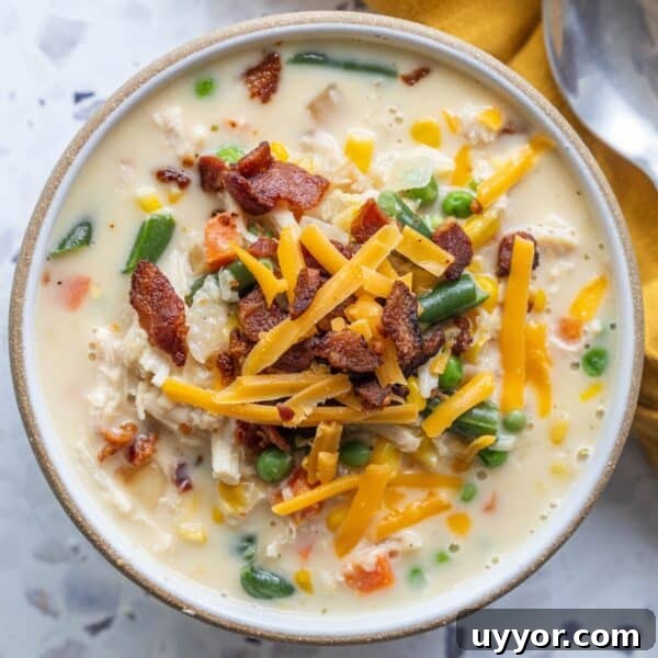 Crispy Honey Mustard Baked Chicken Drumsticks Recipe 15 A bowl of creamy corn chowder topped with shredded cheddar cheese, crispy bacon, and filled with mixed vegetables like peas, green beans, carrots, and corn. A spoon and napkin are nearby.