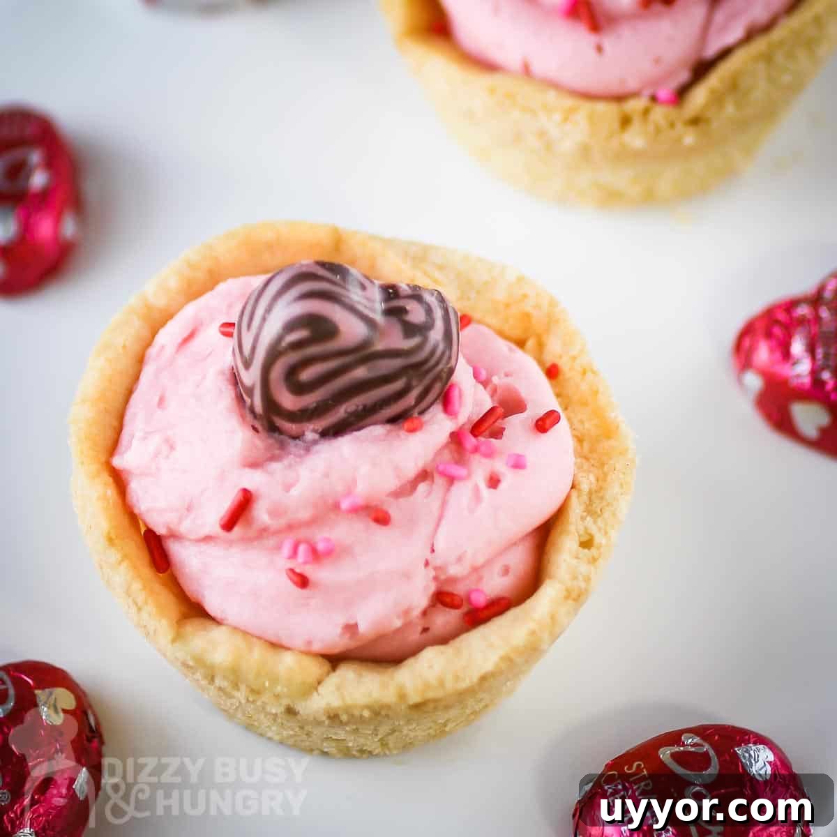 Strawberry Cream Cheese Cookie Cups: Soft Cookies Filled with Creamy Strawberry Centers 6 Close up overhead shot of two cookie cups on a white plate with pink chocolate hearts scattered around.