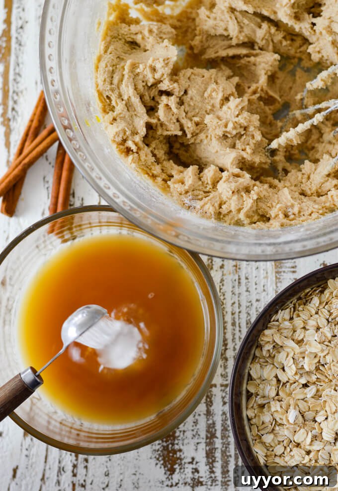 Baking Soda is being poured into raisin water from a measuring spoon. Oatmeal raisin cookie dough sits in a bowl beside it with a bowl of raw oats.