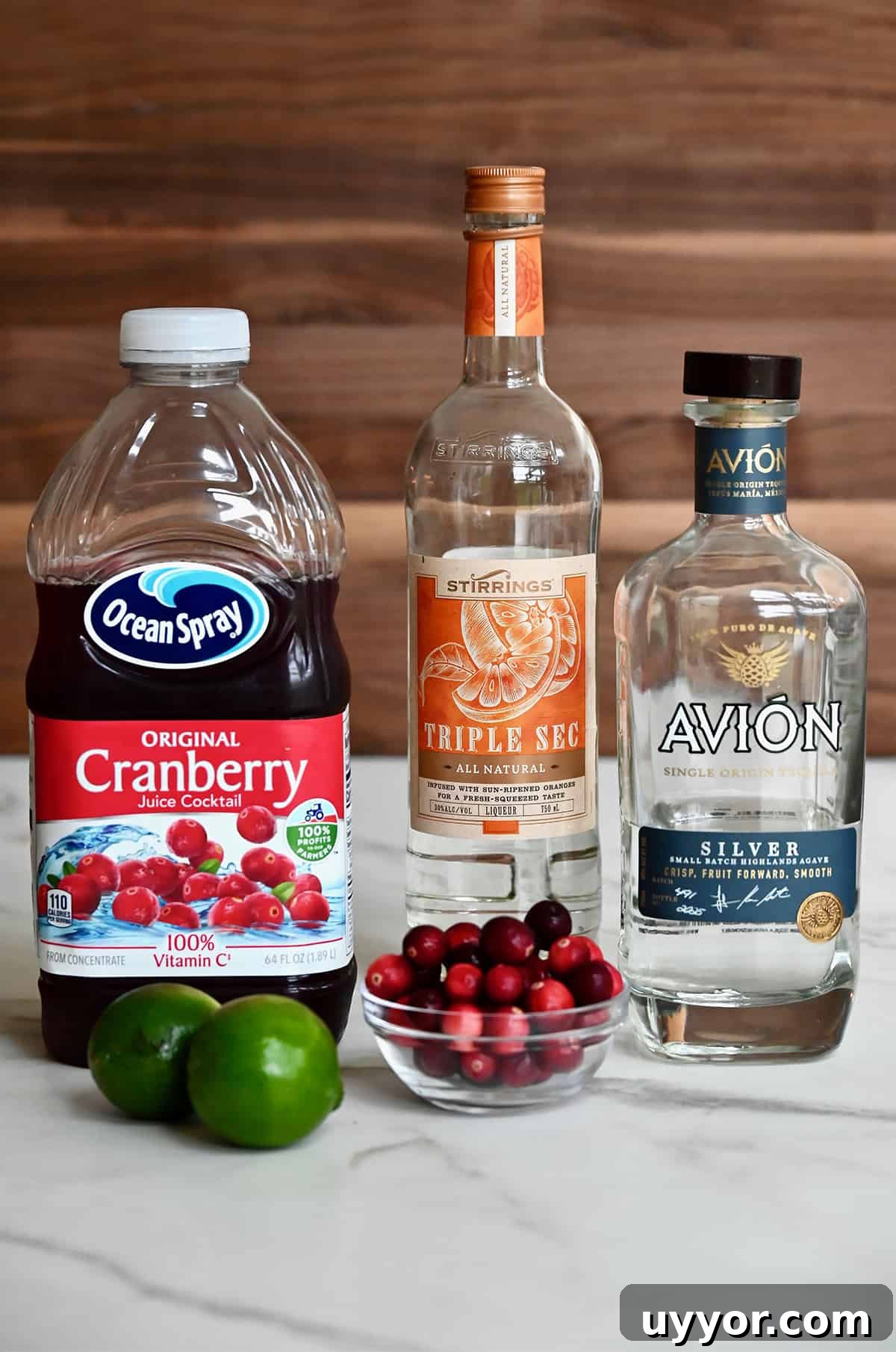 A jug of cranberry juice next to two limes, a bottle of Triple Sec, a bottle of silver tequila and a small bowl filled with fresh cranberries.
