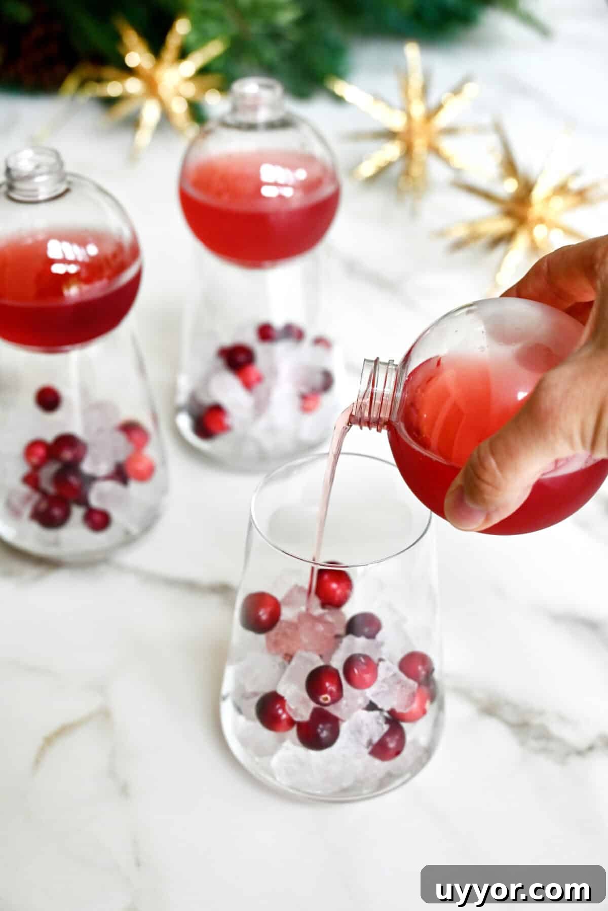 A hand holding a clear plastic ornament drinking ball pours Christmas margarita mix into a glass filled with ice and fresh cranberries.
