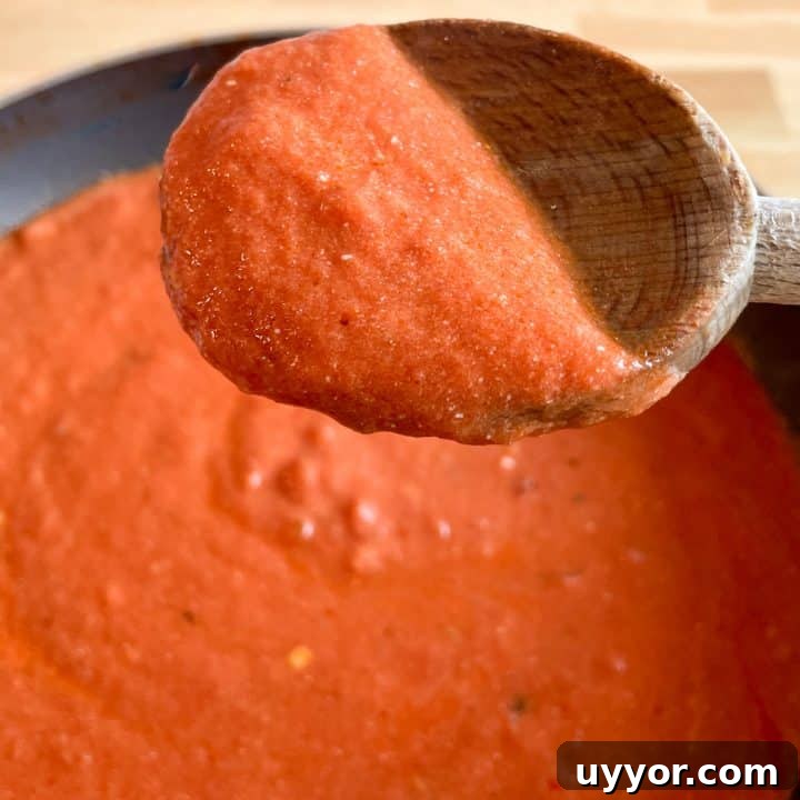 A wooden spoon of tomato and mascarpone sauce is held in the foreground over a pot of cooking sauce in a black pan.