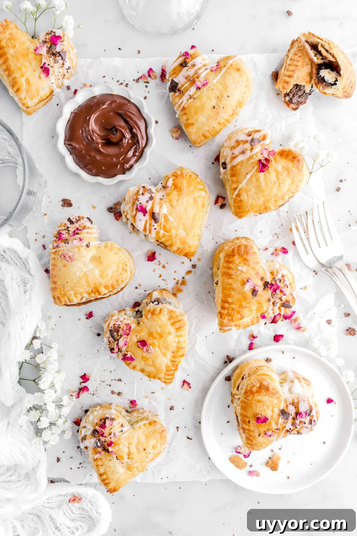 heart shaped puff pastry hand pies on parchment paper with bowl of ganache with dried rose petals and chocolate shavings on each pie.