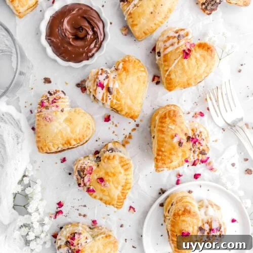 heart shaped puff pastry hand pies on parchment paper with bowl of ganache with dried rose petals and chocolate shavings on each pie.