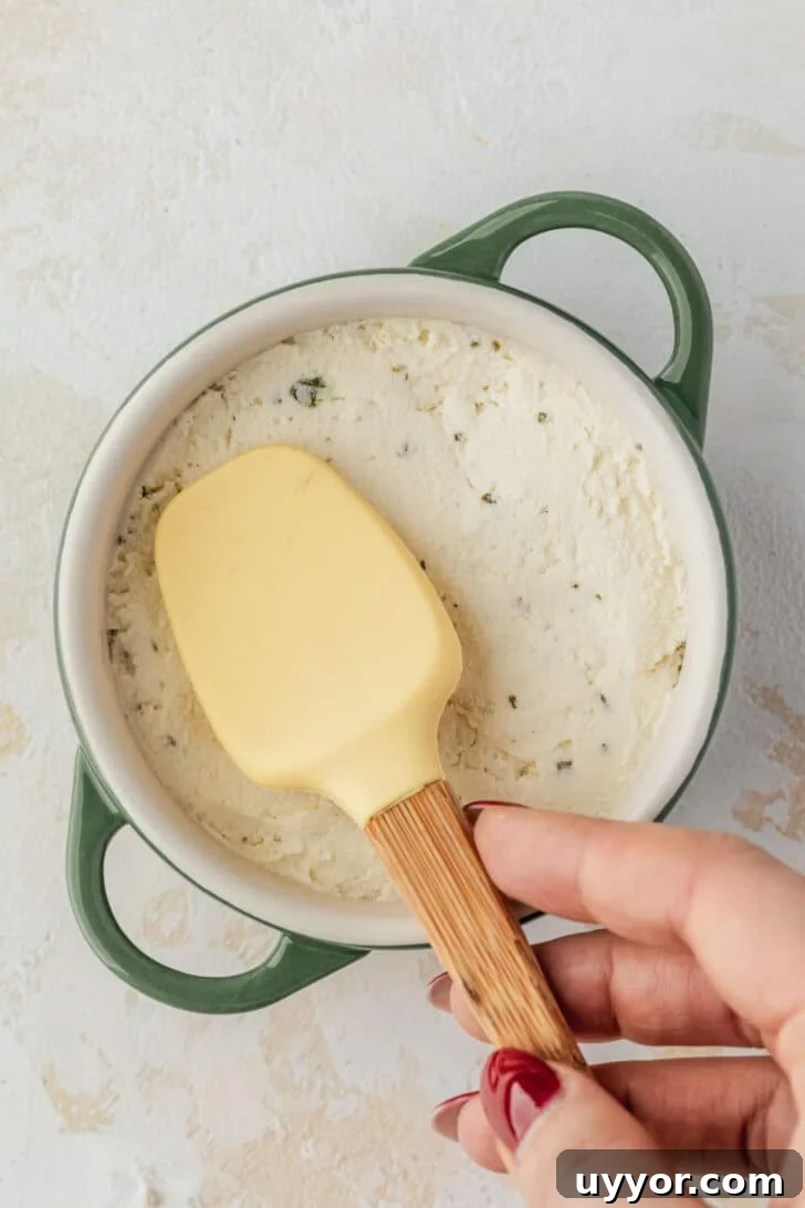 Crispy Baked Boursin Bites Without Dredging 4 A hand using a rubber spatula to spread cheese in a ramekin on a tan counter.