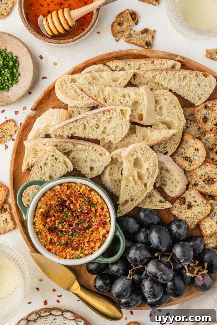 Crispy Baked Boursin Bites Without Dredging 9 A green ramekin of baked boursin on a wood board of bread and grapes next to crackers, glasses of white wine, and brown bowls of chives and hot honey on a tan counter.