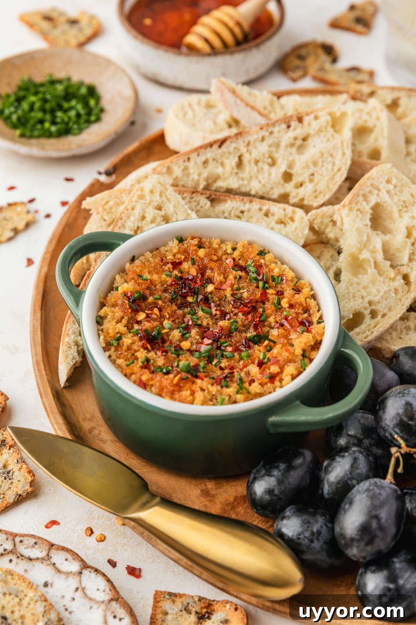 Crispy Baked Boursin Bites Without Dredging 10 A green ramekin of baked boursin cheese with hot honey on a wood board of bread and grapes next to crackers, glasses of white wine, and brown bowls of honey and chives on a beige counter.
