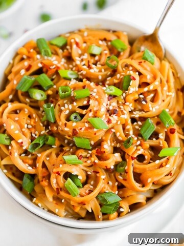 A white bowl filled with creamy peanut butter noodles. They are garnished with green onions and white sesame seeds.