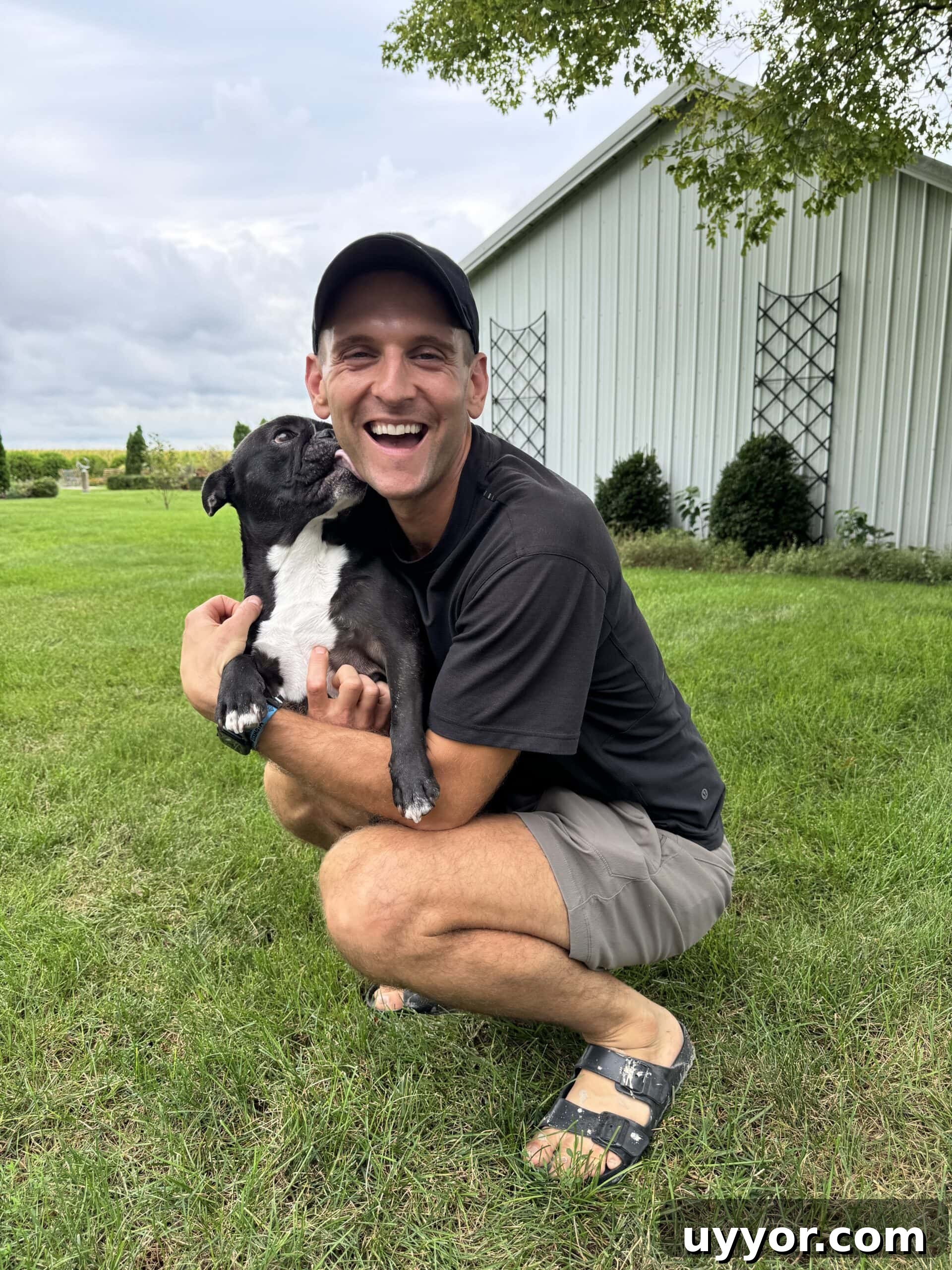 August 2024 Events, Deals & Planning Guide 11 Kaleb crouching in grass with black French bulldog named Kip licking his face.
