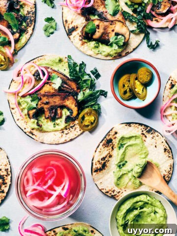 Wide shot of five smoky mushroom tacos on a counter being dressed with toppings like smoky mushrooms, avocado crema, pickled red onions and freshly chopped kale