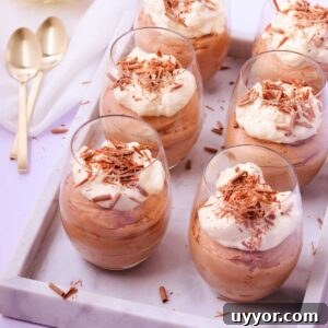 Six stemless wine glasses filled with milk chocolate mousse and topped with whipped cream and chocolate shavings, on a marble tray, with gold teaspoons.