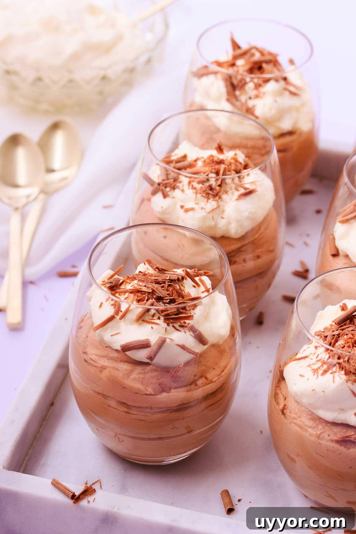 Six stemless wine glasses filled with milk chocolate mousse and topped with whipped cream and chocolate shavings, on an offset marble tray, with gold teaspoons.