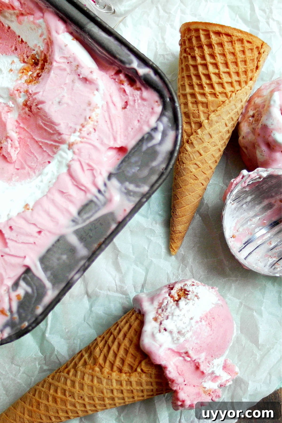 A metal pan of strawberry cheesecake ice cream, an ice cream cone and a serving of the ice cream in a cone.