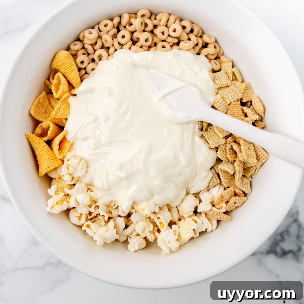 Patriotic Red White & Blue Chex Mix Recipe for 4th of July 5 A white bowl contains popcorn, Chex cereal, Bugles, Cheerios, and a mound of white melted coating being stirred with a white spatula.